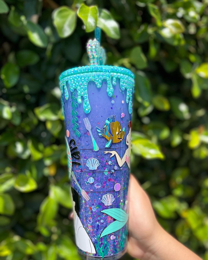 May include: A purple and blue glitter tumbler with a mermaid theme. The tumbler has a blue and green dripping design, a fork and knife, a seashell, and a fish. The tumbler also has a mermaid tail and the text "Ariel" on the side.
