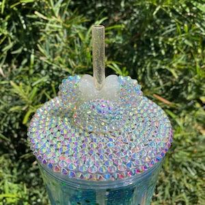 May include: A clear plastic tumbler with a green glitter base and a clear lid. The lid is decorated with iridescent rhinestones and a white plastic Mickey Mouse ear design. A clear plastic straw is inserted through the lid.