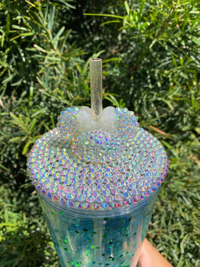 May include: A clear plastic tumbler with a green glitter base and a clear lid. The lid is decorated with iridescent rhinestones and a white plastic Mickey Mouse ear design. A clear plastic straw is inserted through the lid.