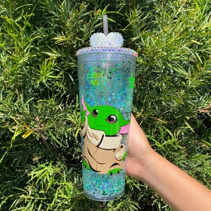 May include: A clear plastic tumbler with a green glitter design and a cartoon image of a green alien character. The tumbler has a clear lid with a rhinestone heart on top.