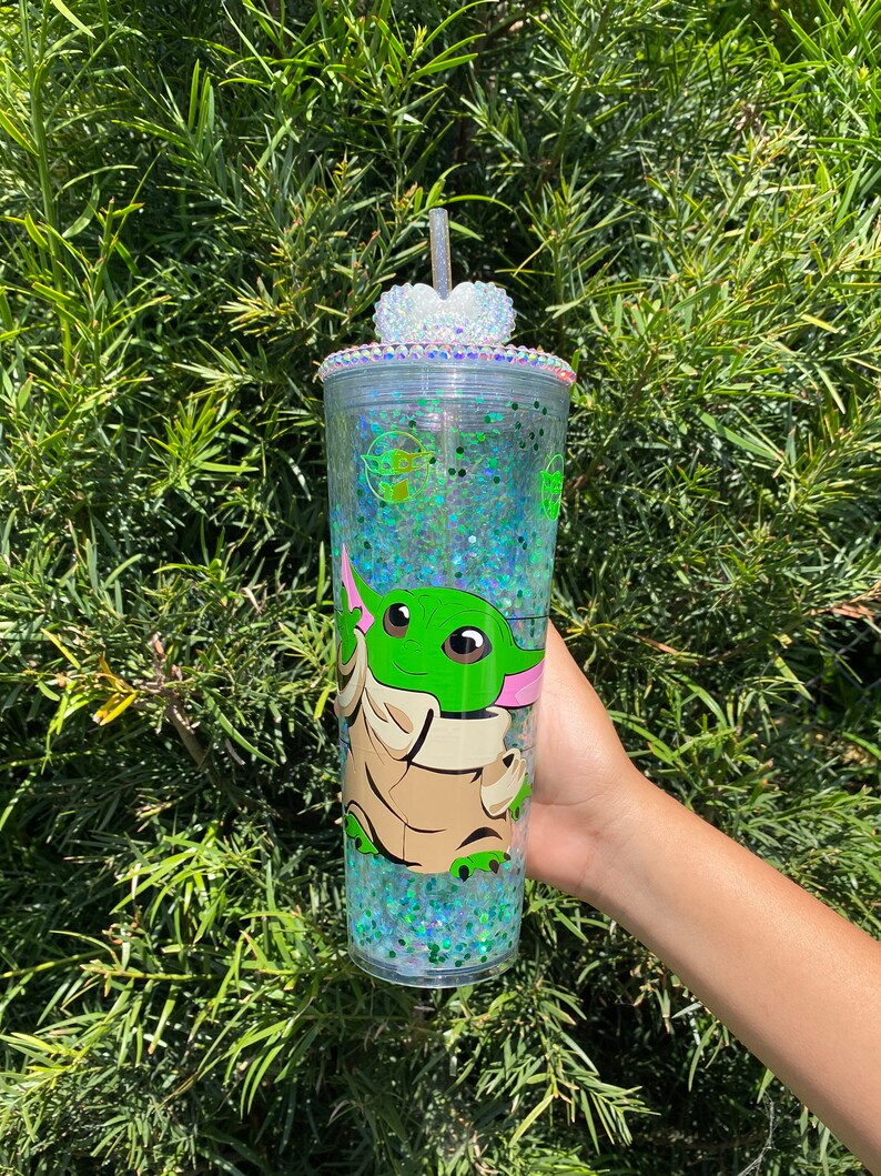 May include: A clear plastic tumbler with a green glitter design and a cartoon image of a green alien character. The tumbler has a clear lid with a rhinestone heart on top.