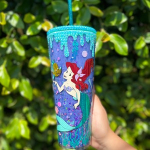 May include: A clear tumbler with a purple glitter base and a mermaid design. The tumbler has a blue and green glitter rim and a blue and green glitter drip effect. The mermaid is holding a seashell and has a red tail. There is a small fish next to the mermaid.
