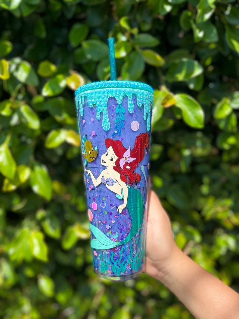 May include: A clear tumbler with a purple glitter base and a mermaid design. The tumbler has a blue and green glitter rim and a blue and green glitter drip effect. The mermaid is holding a seashell and has a red tail. There is a small fish next to the mermaid.