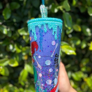 May include: A purple and blue glitter tumbler with a mermaid design and a turquoise drip effect. The tumbler has a straw and is decorated with seashells and forks.
