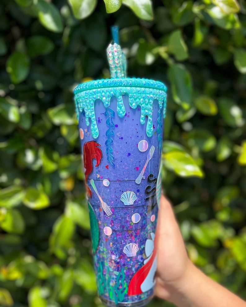 May include: A purple and blue glitter tumbler with a mermaid design and a turquoise drip effect. The tumbler has a straw and is decorated with seashells and forks.