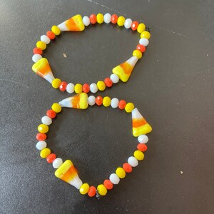 May include: Two candy corn bead bracelets with white, yellow, and orange beads. The bracelets are made with glass beads and are strung on elastic.