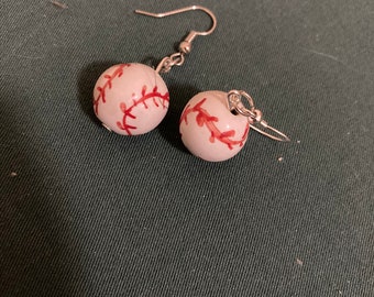 May include: A pair of baseball-themed earrings. Each earring features a small, white ball with red stitching, mimicking a baseball. The earrings have silver-colored hooks.