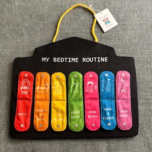 Könnte beinhalten: Eine schwarze Filz-Gute-Nacht-Routine-Tafel mit einer gelben Kordel zum Aufhängen. Sie verfügt über sieben farbige Taschen, die mit Schlafenszeitaktivitäten wie "Potty", "Shower or Bath" und "Good Night" beschriftet sind. Der Text "MY BEDTIME ROUTINE" ist oben aufgedruckt.