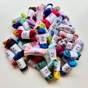May include: A collection of colourful crocheted cords, each wrapped in white paper with the text "Sam Art" printed on it. The cords are arranged in a circle on a white surface.