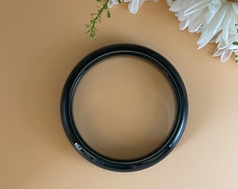 May include: A glossy, dark green jade bangle bracelet. The circular bracelet is set against a neutral background with white flowers in the upper right corner. The bangle is a solid, smooth band.