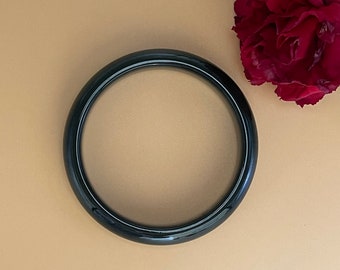 May include: A glossy, dark green bangle bracelet. The circular bracelet is set against a neutral background, with a deep red flower and greenery in the upper right corner. The bracelet's smooth surface reflects light.