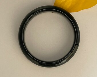 May include: A glossy, dark green bangle bracelet. The circular bracelet is set against a neutral background with a yellow leaf. The bracelet's smooth surface reflects light, highlighting its polished finish. A simple, elegant piece of jewelry.