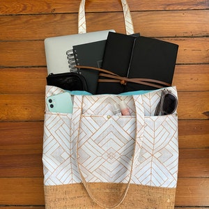 May include: A white tote bag with a cork bottom and a geometric pattern. The bag is filled with various items, including a laptop, notebooks, a phone, and a makeup bag.