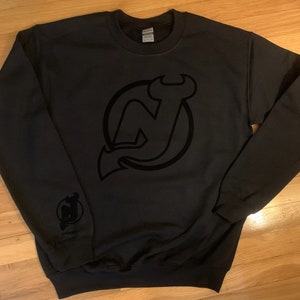 May include: Black sweatshirt with a black New Jersey Devils logo on the front. The logo is a stylized "NJ" with a devil's tail.