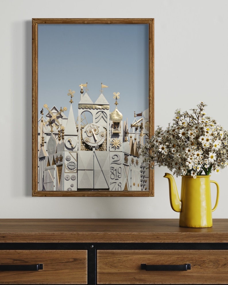K&ouml;nnte beinhalten: Ein gerahmtes Foto eines wei&szlig;en und goldenen Geb&auml;udes mit einem Zifferblatt und filigranen Details. Das Geb&auml;ude steht vor einem blauen Himmel. Das Foto ist auf einem Holzregal mit einer gelben Teekanne und einem Strau&szlig; wei&szlig;er Blumen ausgestellt.