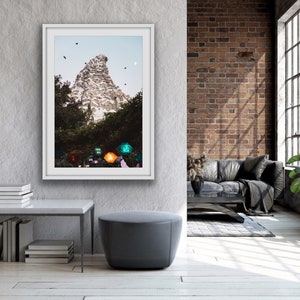 Pu&ograve; includere: Una fotografia incorniciata di una montagna con una cima bianca e alberi verdi in primo piano. La montagna &egrave; circondata da un cielo blu con alcune nuvole bianche. La fotografia &egrave; appesa a una parete grigia in un soggiorno moderno.
