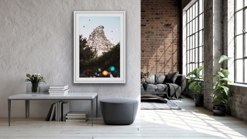 Pu&ograve; includere: Una fotografia incorniciata di una montagna con una cima bianca e alberi verdi in primo piano. La montagna &egrave; circondata da un cielo blu con alcune nuvole bianche. La fotografia &egrave; appesa a una parete grigia in un soggiorno moderno.