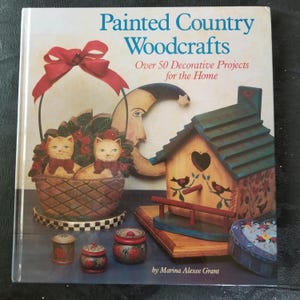May include: Book cover featuring a painted wooden birdhouse, a basket with two kittens, and other painted wooden crafts. The book title is "Painted Country Woodcrafts" with the subtitle "Over 50 Decorative Projects for the Home" by Marina Alexee Grant.