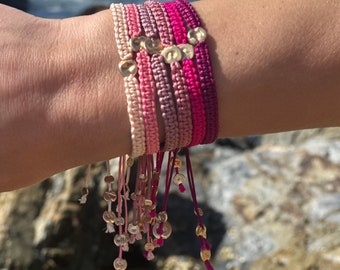 May include: A stack of five colorful friendship bracelets with gold heart charms. The bracelets are made of thin, braided cords in shades of pink, red, and white. The charms are attached to the ends of the bracelets, creating a cascading effect.