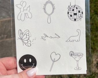 May include: A sheet of temporary tattoos featuring a range of designs, including a balloon animal, a mirror, a disco ball, a butterfly, a dinosaur, a smiley face, a heart-shaped lollipop, and a margarita glass. The sheet also includes the word "lucky" written in cursive.