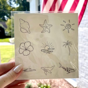 May include: A sheet of temporary tattoos with nine different designs, including a seashell, starfish, sun, hibiscus flower, sunrise, palm tree, wave, whale, and shark. The tattoos are black and white.
