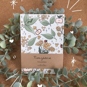 May include: A white notebook with a floral pattern featuring red ladybugs, green leaves, and yellow flowers. The notebook has a brown paper label that reads "Notizblock" with the text "Wiesenleben" and "Himmelstau Creation" underneath.