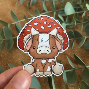 May include: A cartoon cow wearing a red and white mushroom hat. The cow is brown with white spots and has a heart-shaped spot on its side. The mushroom has white dots on a red background.