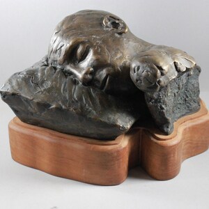 May include: Bronze sculpture of a person sleeping on a pillow. The sculpture is on a wooden base.