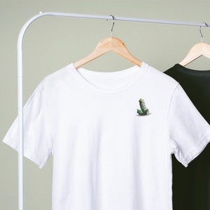 Könnte beinhalten: Zwei kurzärmelige T-Shirts, eines weiß und eines schwarz, hängen an einer Kleiderstange. Jedes Shirt hat eine kleine, grüne Grafik auf der oberen linken Brust. Die Shirts sind aus weichem Material und vor hellgrünem Hintergrund.
