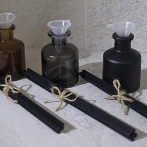 May include: Three glass diffuser bottles in brown, grey, and black, each with a clear plastic funnel. Bundles of black diffuser sticks are tied with twine. The bottles are for aromatherapy or home fragrance.