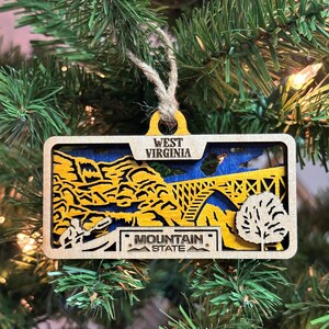 May include: Wooden Christmas ornament shaped like a West Virginia license plate with a blue and yellow mountain scene and the text "West Virginia Mountain State".