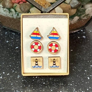 May include: A set of six wooden earrings in a small box. The earrings feature nautical themes, including sailboats, life preservers, and lighthouses. The sailboats are red, yellow, and blue. The life preservers are red and white. The lighthouses are black and white.