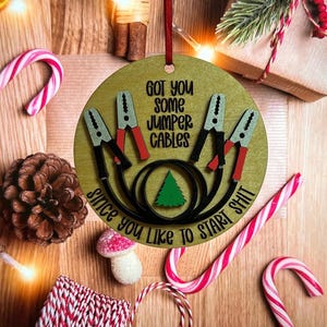 May include: A round, gold-colored Christmas ornament with the text "Got you some jumper cables since you like to start shit". The ornament features a black and white illustration of jumper cables and a small green Christmas tree.