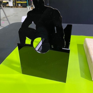 May include: Black silhouette of a person in a crouching pose, cut out of acrylic and mounted on a black acrylic base. The base has a circular opening in the center.