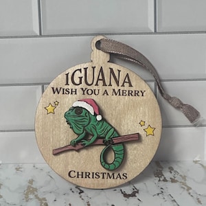 May include: A wooden Christmas ornament shaped like a round ball with a green iguana wearing a red Santa hat on a branch. The ornament has the text "Iguana Wish You a Merry Christmas" on it.