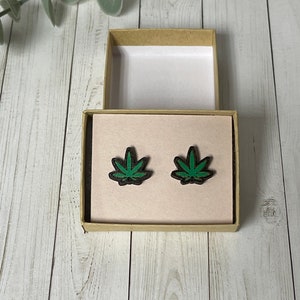 May include: A pair of green marijuana leaf stud earrings in a small cardboard box with a white interior.