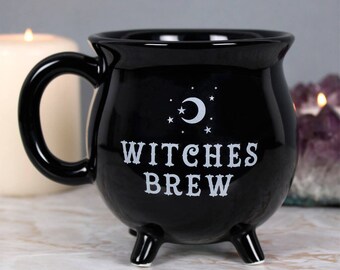 May include: Black ceramic mug shaped like a cauldron with a white crescent moon and stars design. The mug has a handle and three legs. The text "Witches Brew" is printed on the front.