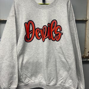 May include: A light gray crewneck sweatshirt featuring the word Devils printed in a bold, red script font with a thick black outline. The garment has long sleeves, a ribbed collar, and a standard fit, displayed on a bright green plastic hanger.