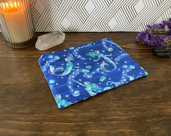 May include: A blue fabric pouch with a mermaid and star pattern. The pouch is on a wooden surface.