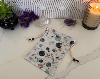 May include: A white drawstring bag with a celestial print featuring constellations, moons, and stars. The bag has black beads on the drawstring.