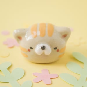 May include: A small ceramic figurine of a cat with orange stripes and a white face. The cat has black eyes and a small black nose. The figurine is sitting on a yellow background with green leaves and pink flowers.