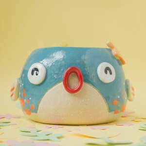 May include: A blue and white ceramic planter shaped like a pufferfish with a red mouth and black eyes. The planter has a white belly and orange spots.
