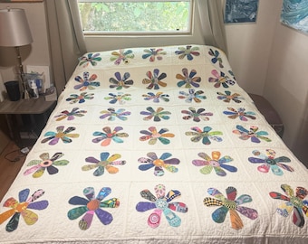 May include: A cream-colored quilt with a repeating pattern of large, colorful flower appliqués. Each flower features a variety of patterned fabrics in shades of blue, purple, orange, and green. The quilt is on a bed near a window.