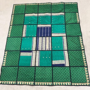 May include: A patchwork quilt with a geometric design. The quilt features squares of teal, navy, and patterned green fabric, outlined with black stitching. The border is green with a gold trim and small tassels.