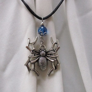 May include: A silver spider pendant necklace with a blue glass bead. The spider has a detailed web design and is hanging from a black cord.
