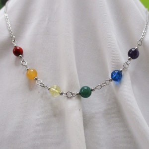 May include: A silver chain necklace with seven round gemstone beads in the colors of the rainbow. The beads are red, orange, yellow, green, blue, indigo, and violet.