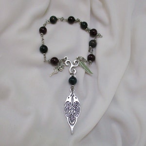 Peut inclure: Un bracelet de perles avec une chaîne argentée et des breloques décoratives. Le bracelet est composé de perles rouge foncé et vertes. Un pendentif argenté au design complexe est suspendu, ainsi qu'une plume et d'autres breloques.