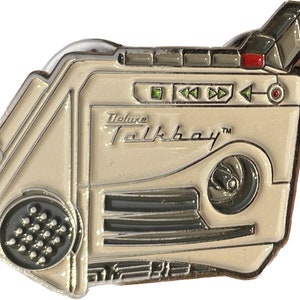 Może przedstawiać: Biała emaliowana przypinka przedstawiająca odtwarzacz kasetowy w stylu vintage z napisem "Deluxe Talkboy™" z przodu. Przypinka ma srebrne wykończenie i zawiera mikrofon, głośnik oraz przyciski sterujące.