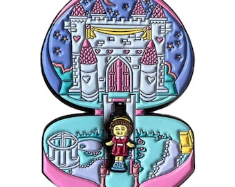 Puede incluir: Un pin de esmalte rosa y azul con forma de corazón. El pin está abierto para revelar un castillo en miniatura con una muñeca dentro. El castillo es blanco con detalles en rosa y azul. La muñeca lleva un vestido rojo y tiene el pelo castaño.