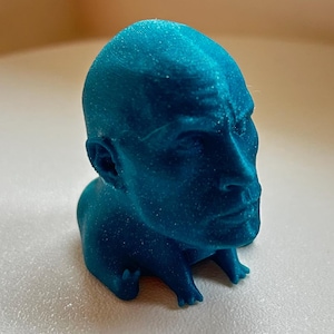 Könnte beinhalten: Eine türkisfarbene Skulptur eines menschlichen Kopfes mit detaillierten Gesichtszügen. Die kleine Skulptur scheint aus einem strukturierten Material zu bestehen. Der Kopf ist aufrecht, Hals und Schultern bilden eine kleine Basis.